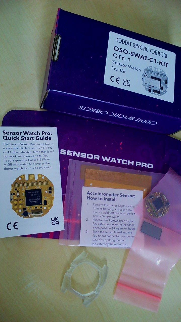 Picture of the coloreful box of the Sensor Watch Pro by Oddly Specific Objects, its content and a 3d printed watch body (clear resin).