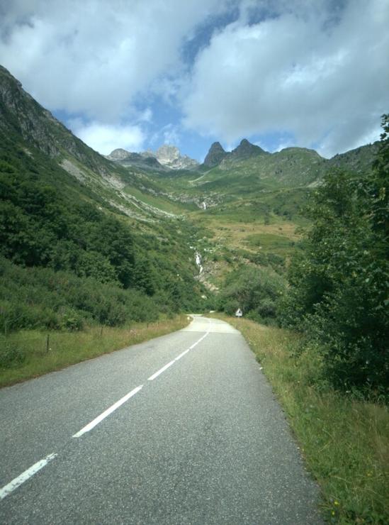 road leading downhill towards a stream with some cascades, rocky mountains in the background